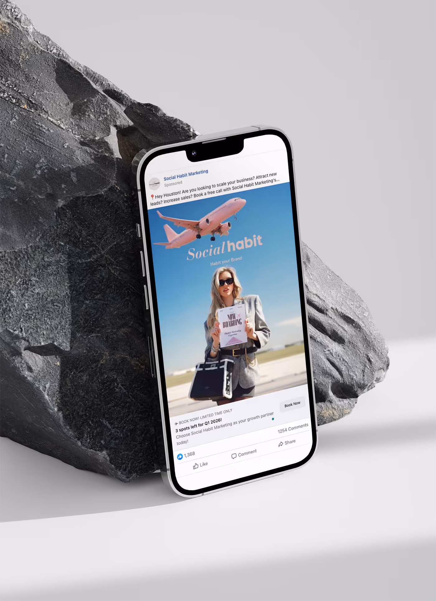 Smartphone leaning against a grey rock displaying a Social Habit Marketing ad featuring a woman holding a 'Now Boarding' sign with a pink airplane in the background.